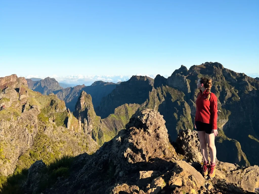 Girl in a red patagonia top, black cycling shorts and hiking boots at the famous pico to pico hike in Madeira looking out over the surrounding mountains at sunrise.