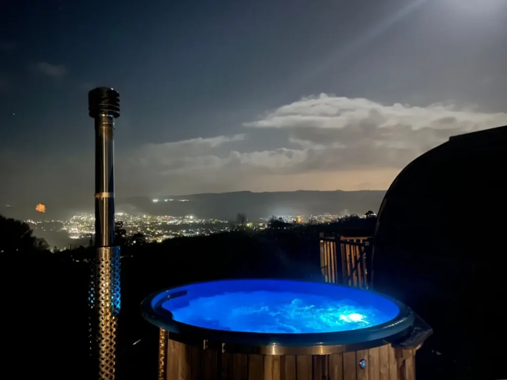 Juniper cabin hot tub set high on the hill overlooking nearby village at Stoneymollan over Loch Lomond at night.