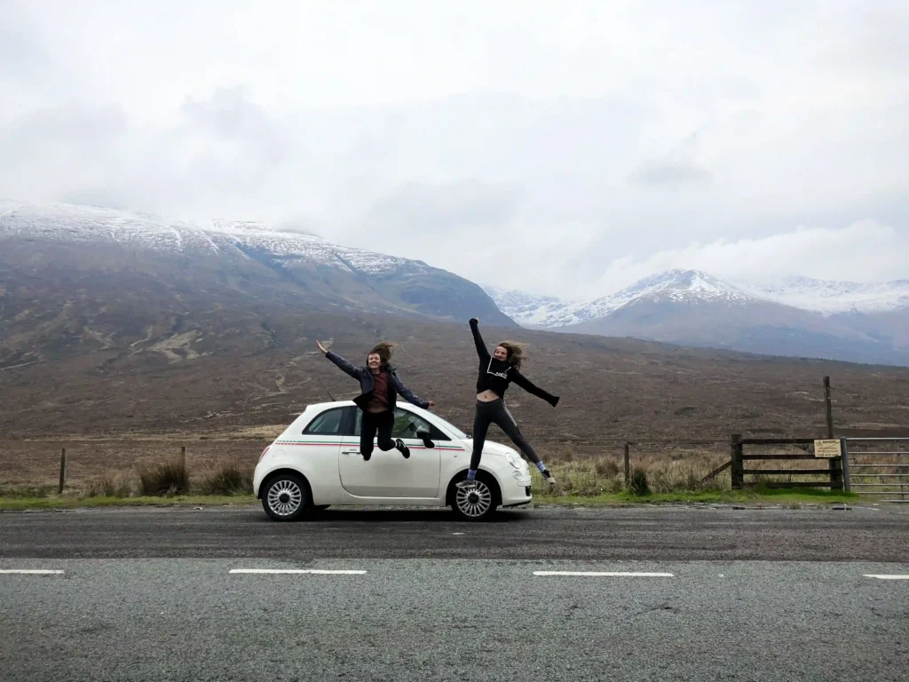 Two girls jumping in front of their white fiat 500 car with the scottish highlands in the background.