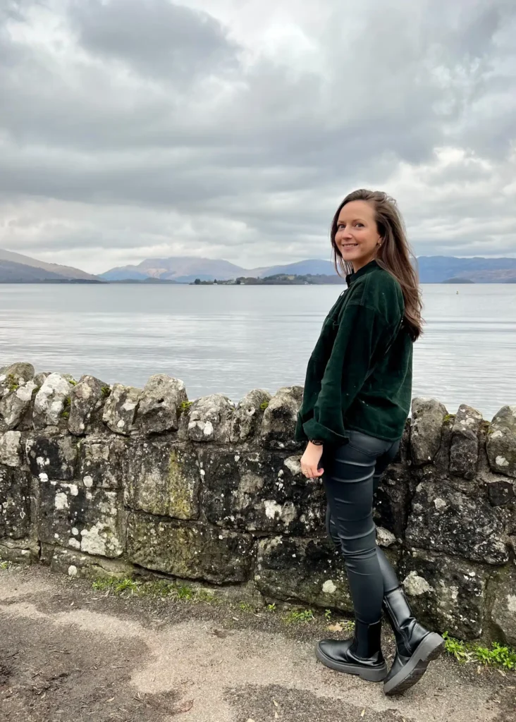 Girl standing smiling at the camera with Loch Lomond in the background on a cloudy day.