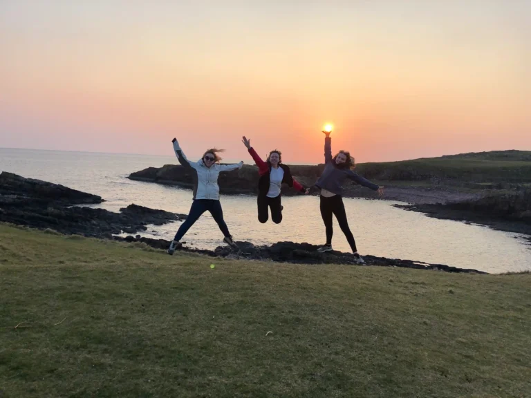 Three girls jumping for joy while the sun sets at Clachtoll beach on the North Coast 500 in Scotland.