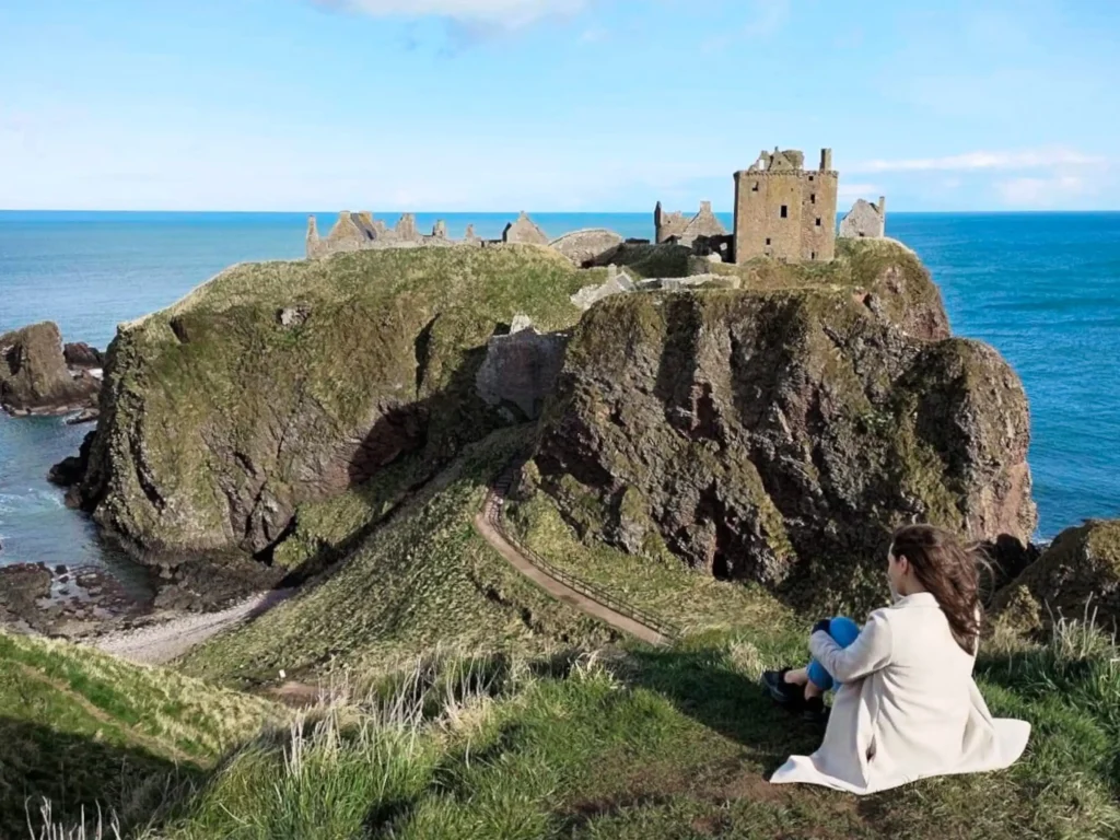 Girl sitting down looking windswept and over Dunnottar Castle in Scotland on a sunny day.