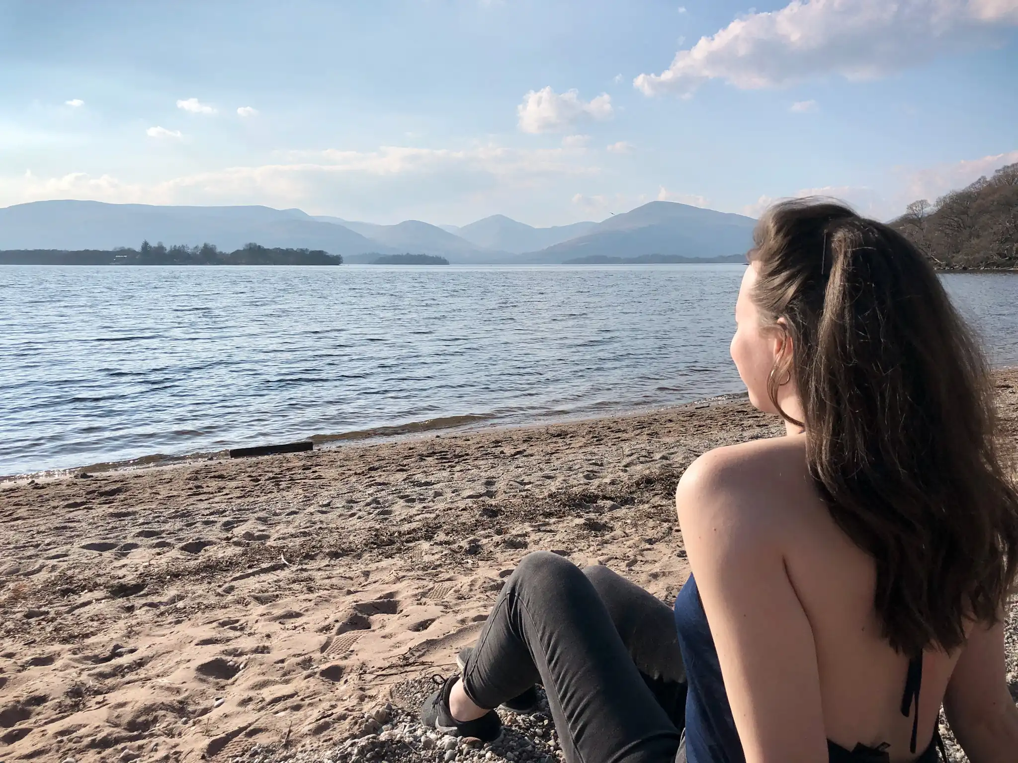 Girl sitting with her back to the camera on Milarrochy Bay near Balmaha in Scotland overlooking Loch Lomond.