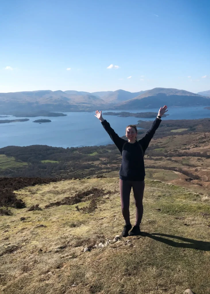 Girl with hands up in the air looking happy at the summit of Conic Hill in Loch Lomond, Scotland.
