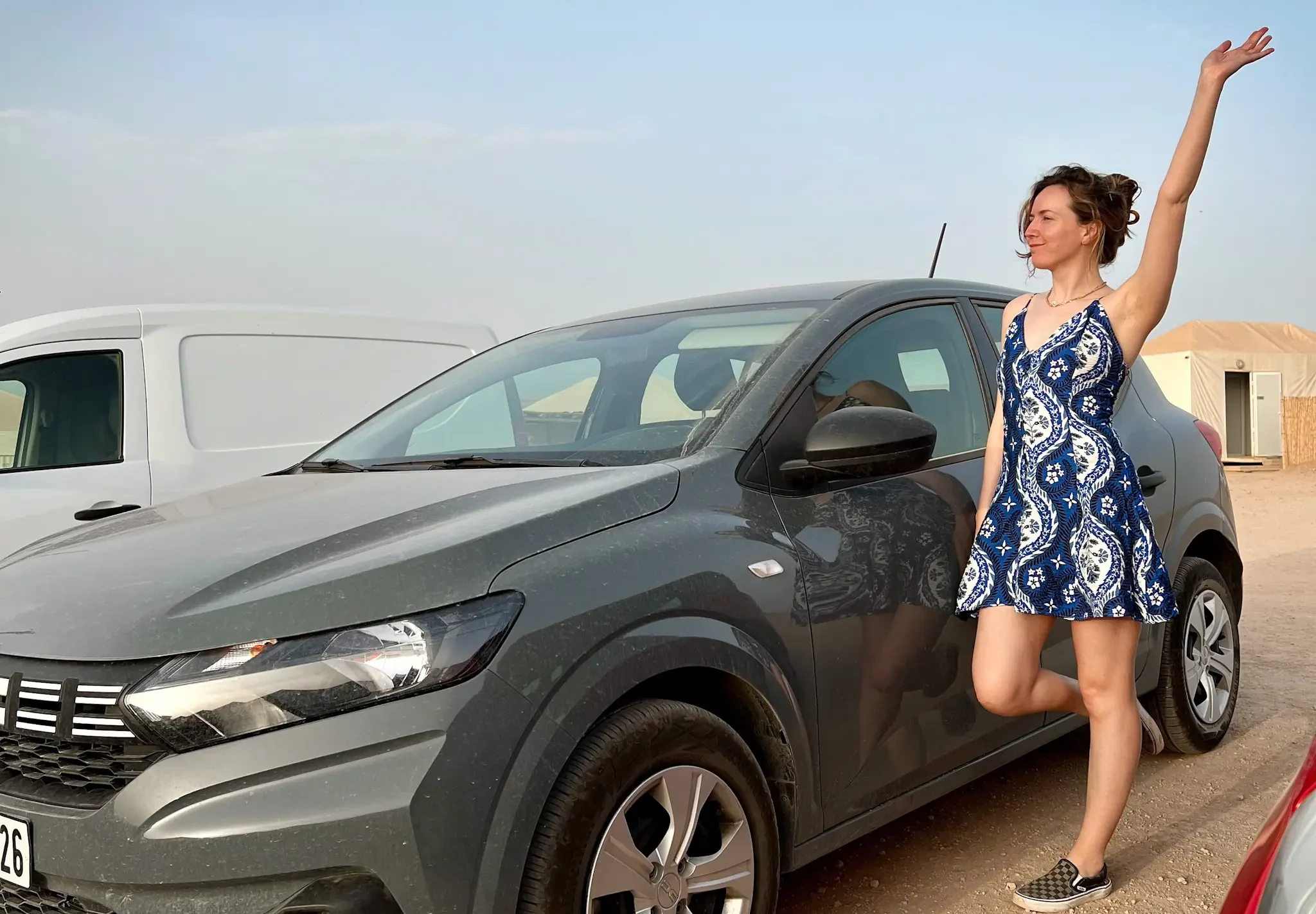 Girl standing next to grey Dacia Sandero with hand in the air.