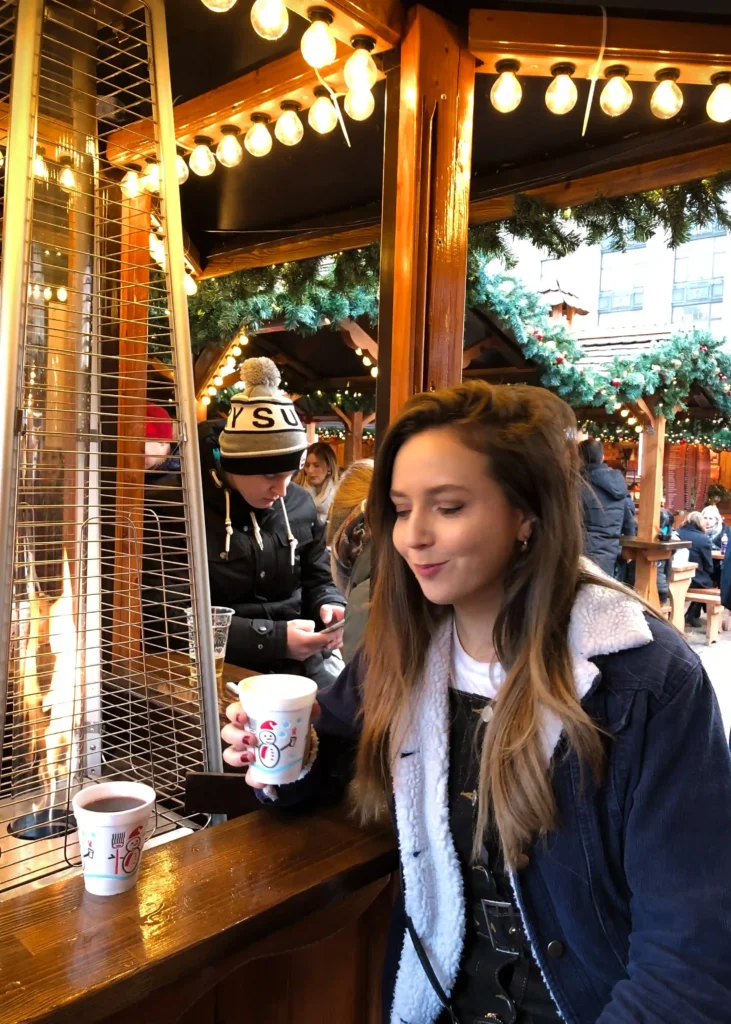 Girl smiling looking at her mulled wine while sitting next to a warm fire at a Christmas market.