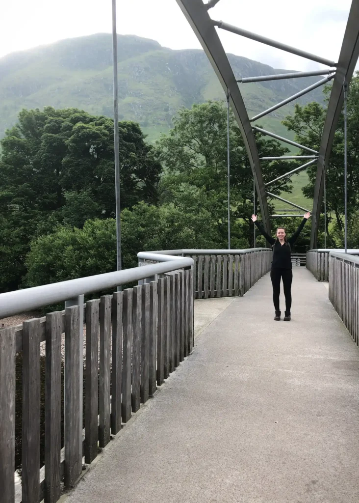 Girl standing on a bridge with arms in the air and Ben Nevis mountain range in the background.