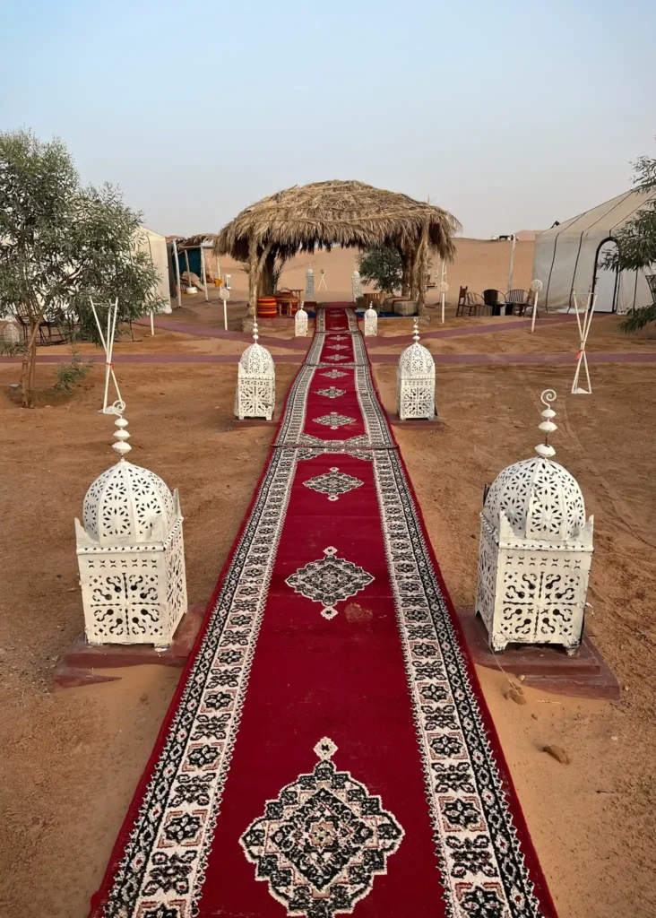 Desert camp in Merzouga with huts, lanterns, and rugs.