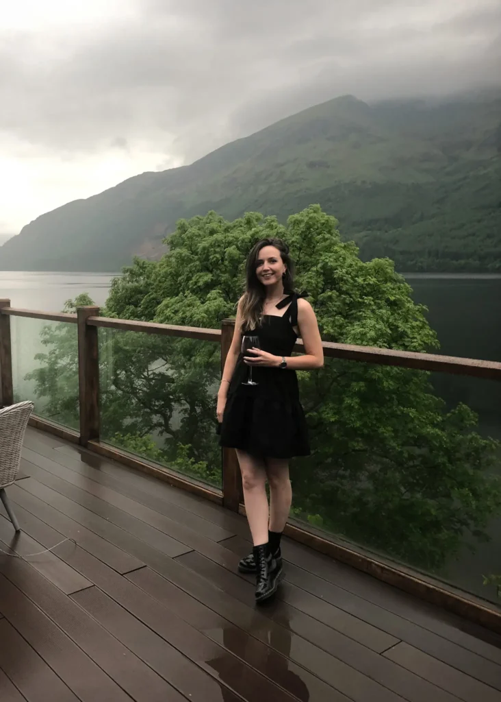 Girl standing holding a glass of wine smiling at the camera on decking with a loch and hills behind her.