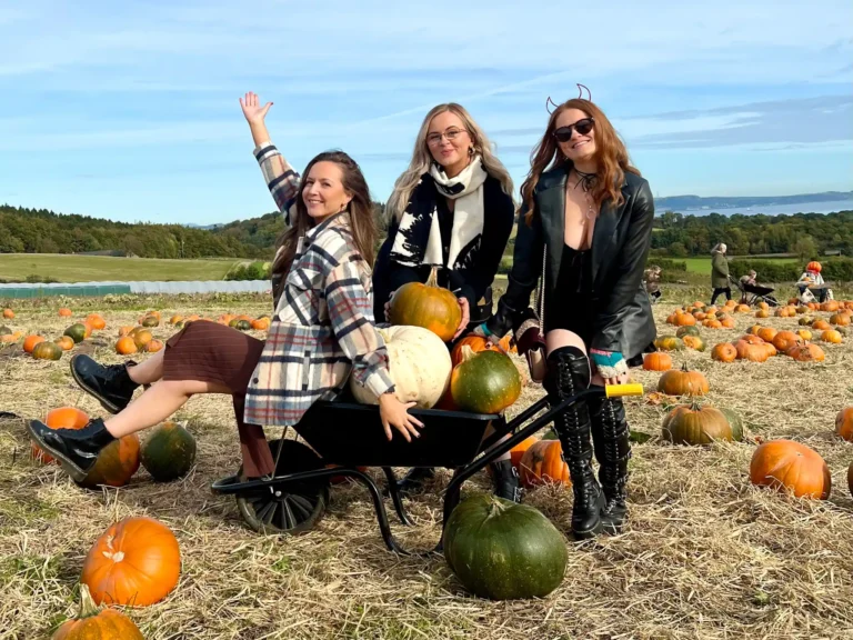Three girls smiling at the camera sitting on a wheelbarrow at a pumpkin patch in Scotland.