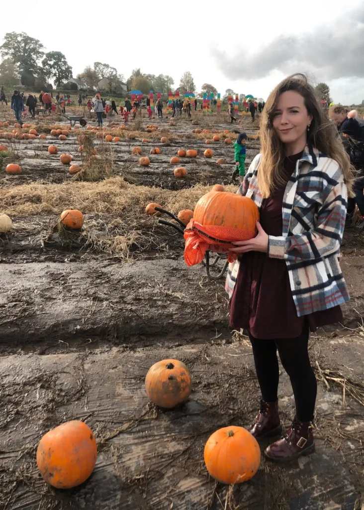 Girl holding a pumpkin looking at the camera with a muddy pumpkin patch behind her.