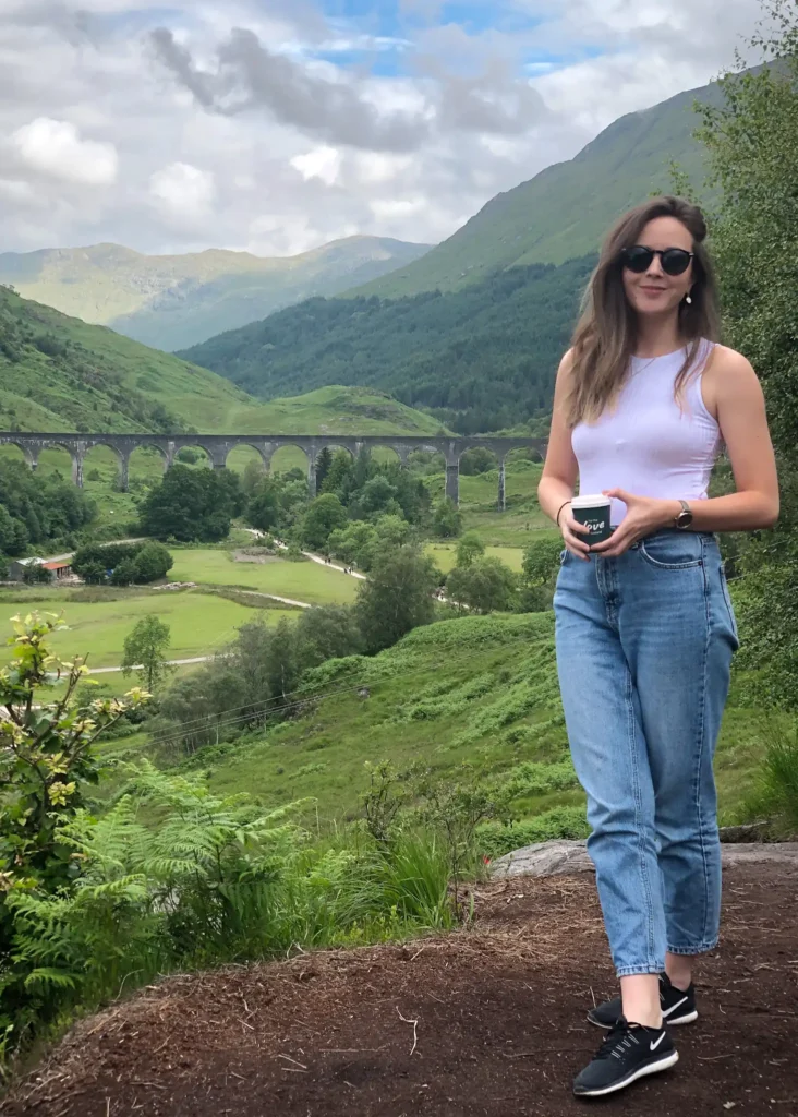 Girl standing smiling at the camera holding a coffee cup with the Glenfinnan Viaduct in the background.