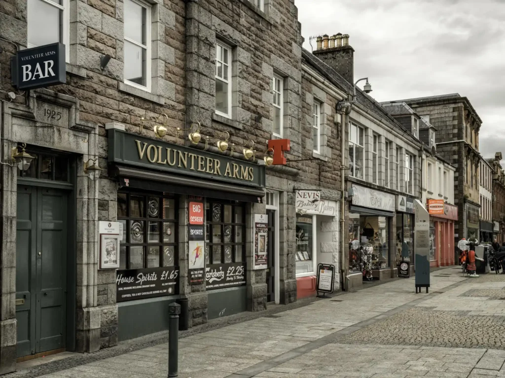 Street in Fort William lined with bars, restaurants, and shops on a cloudy day.