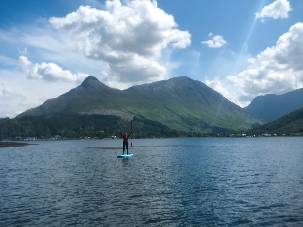 Girl standing on a paddleboard in a loch in Glencoe with hills in the background.