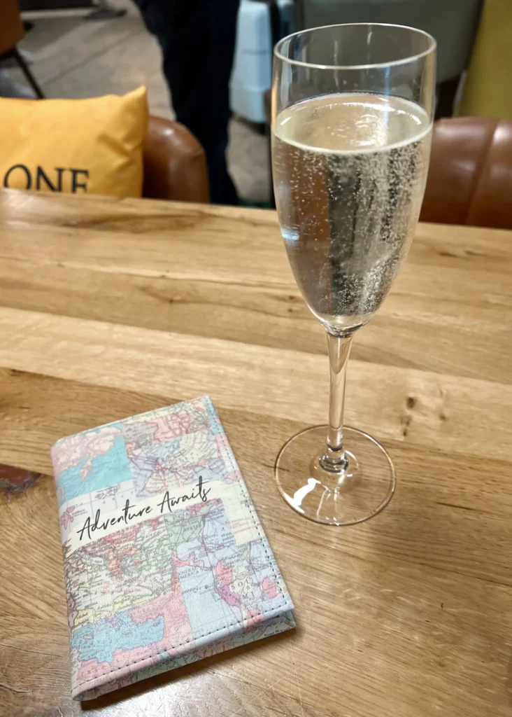 Passport with world-map cover saying "adventure awaits" with a glass of prosecco.