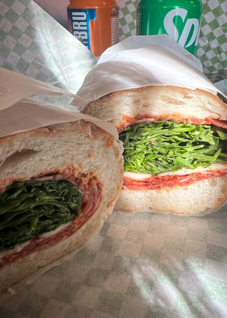 Baguette cut in two showing the fillings of salami, rocket, cheddar, hot honey and sriracha butter with cans of fizzy drinks in the background