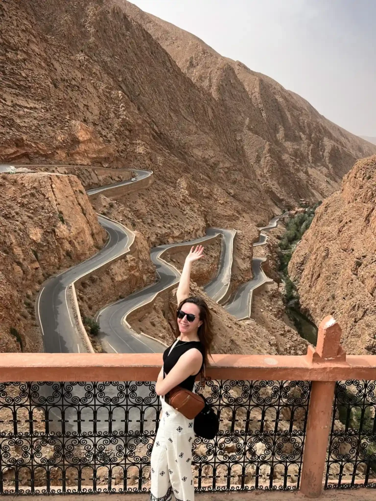 Girl standing at a viewpoint overlooking Tisderine Bends in Dades Valley with one hand up and smiling at the camera.
