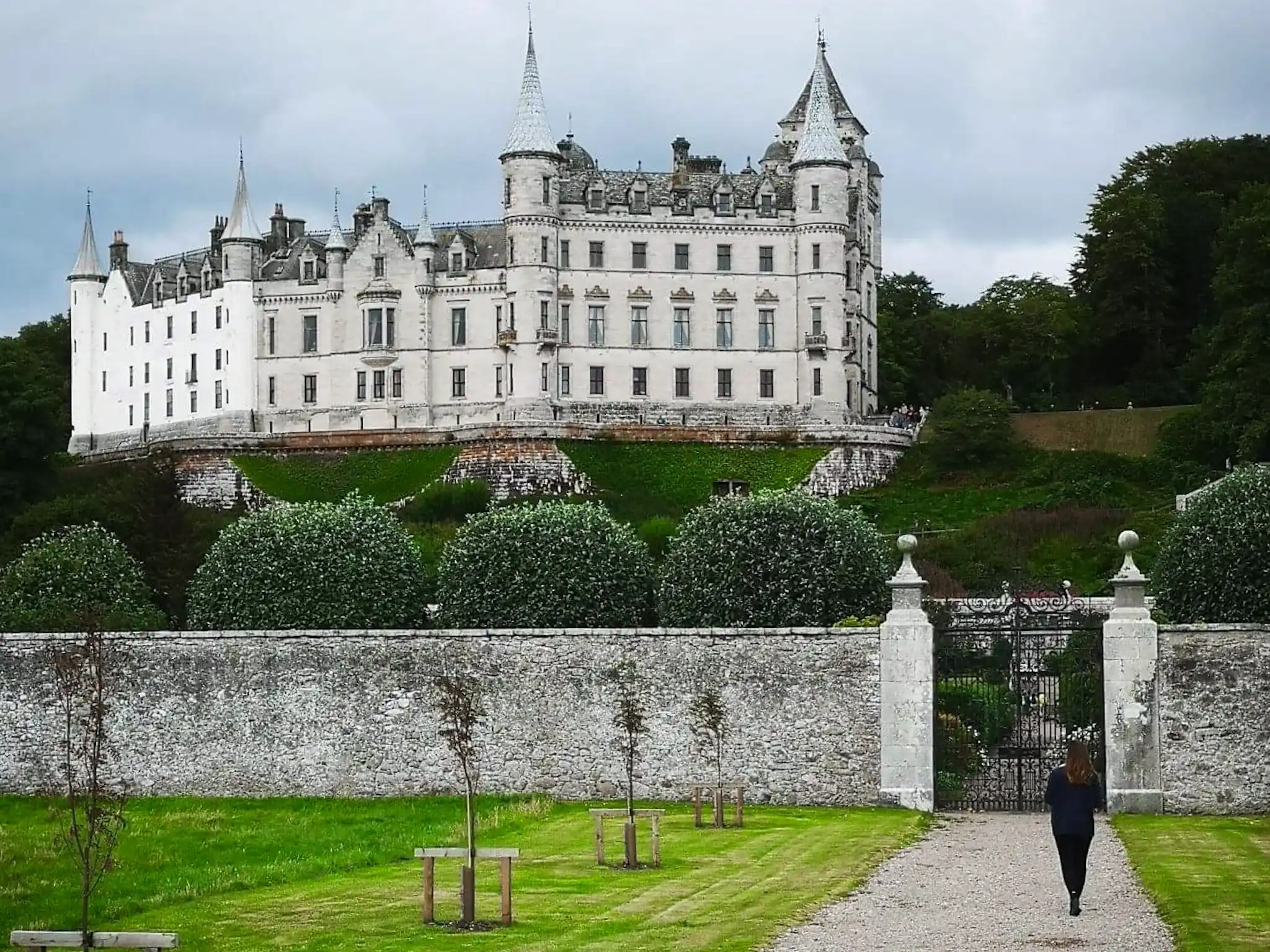 Girl walking towards a walled gate with Dunrobin Castle in the background on a cloudy day.