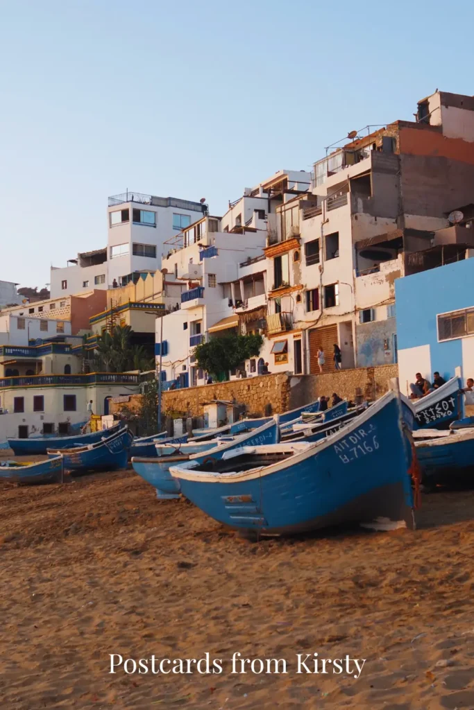 Pinterest Pin showing Taghazout beach with boats on the sand and the buildings and town in the background at sunset with "Postcards from Kirsty" text.