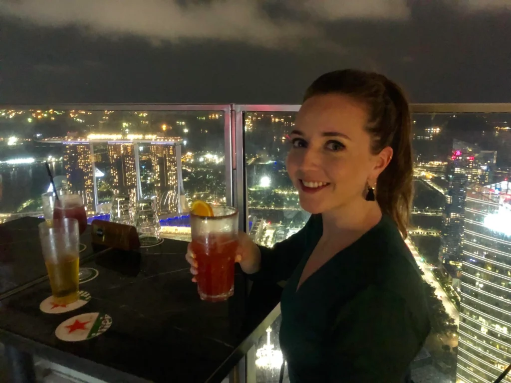 Woman smiling at camera holding a cocktail on a Singapore rooftop bar overlooking the skyline.