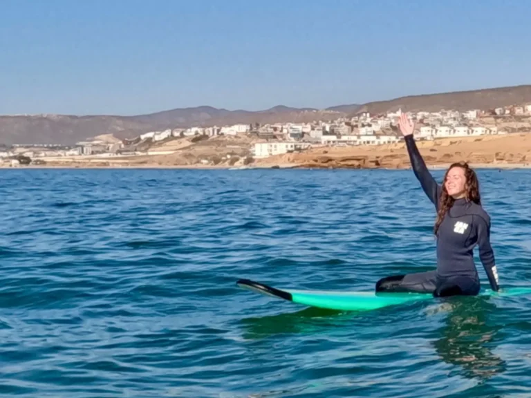 Girl sitting on her surfboard with hands up in the air, smiling in the water at Taghazout.