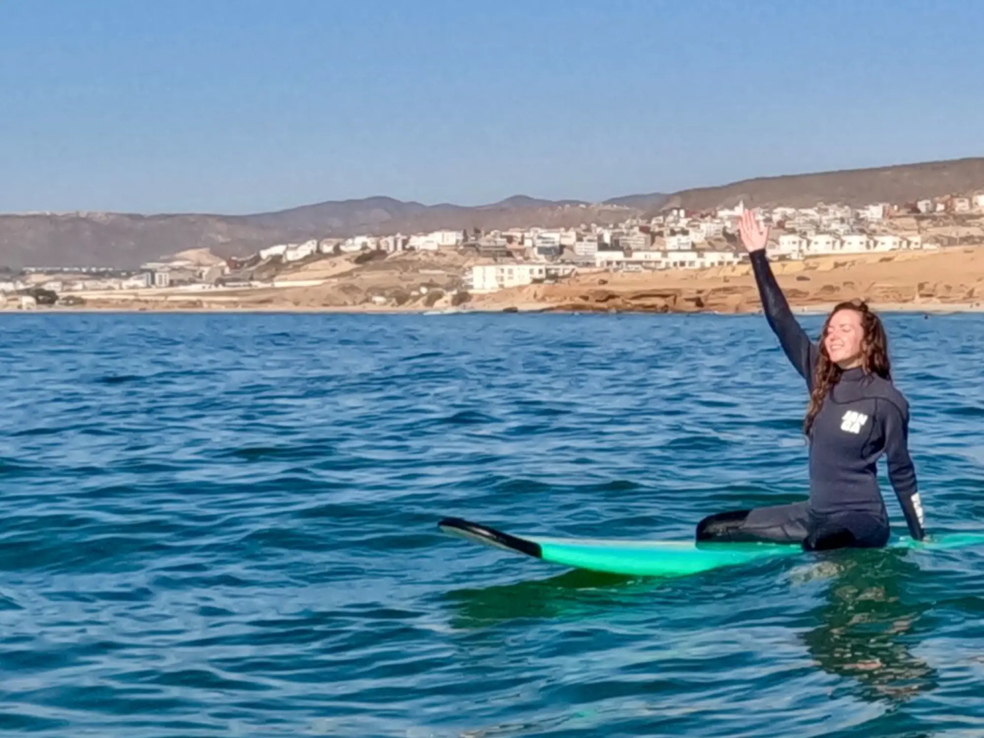 Girl sitting on her surfboard with hands up in the air, smiling in the water at Taghazout.
