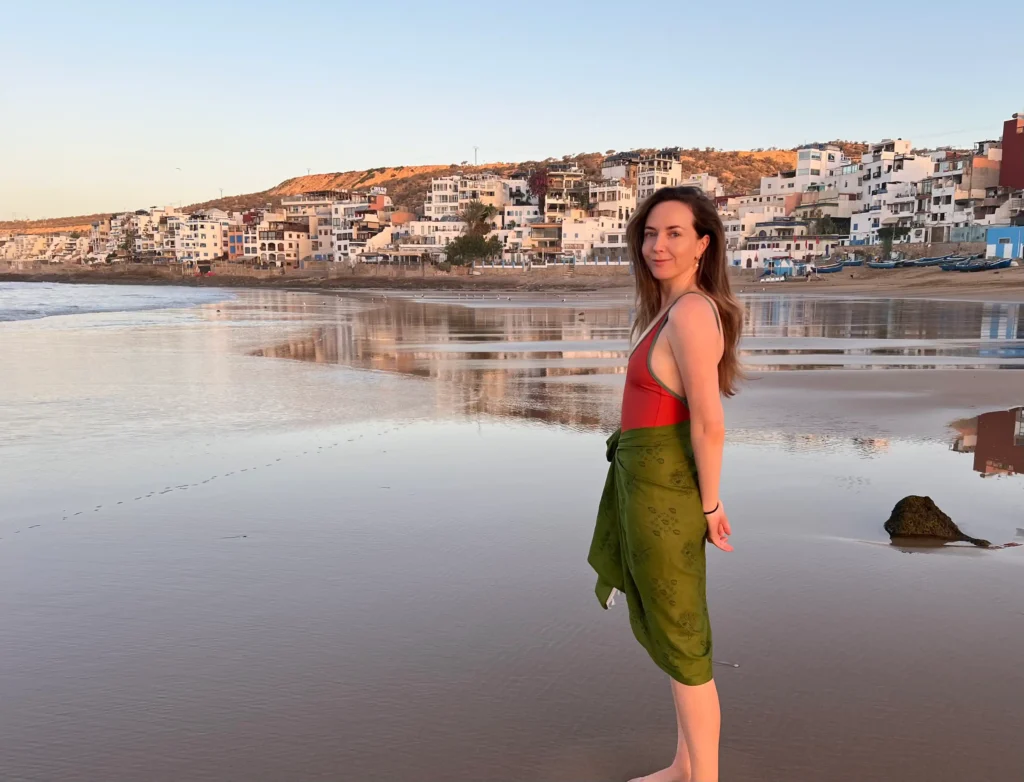 Girl standing side on to the camera in a red swimsuit and green sarong, smiling with Taghazout in the background at sunrise.