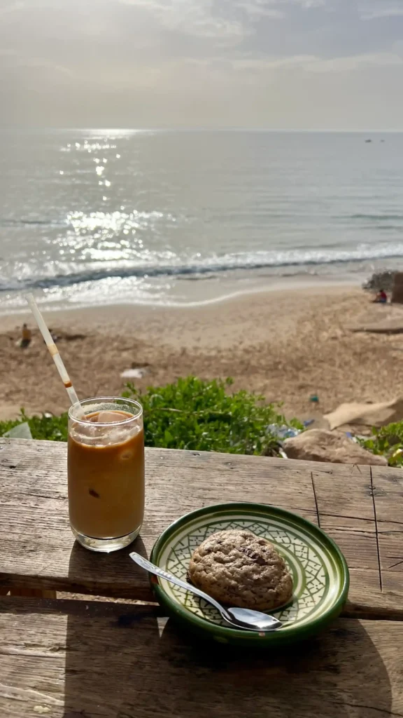 An iced latte with a Nutella cookie on a table with the beach and ocean in the background.