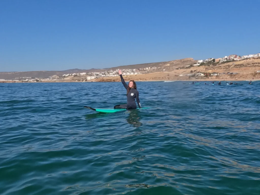 Girl sitting on her surfboard with hands up in the air, smiling in the water at Taghazout.