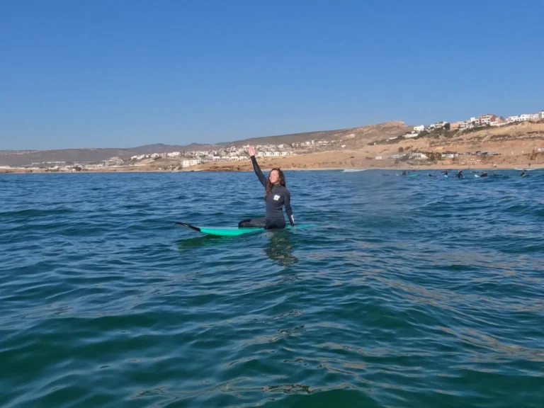 Girl sitting on her surfboard with hands up in the air, smiling in the water at Taghazout.