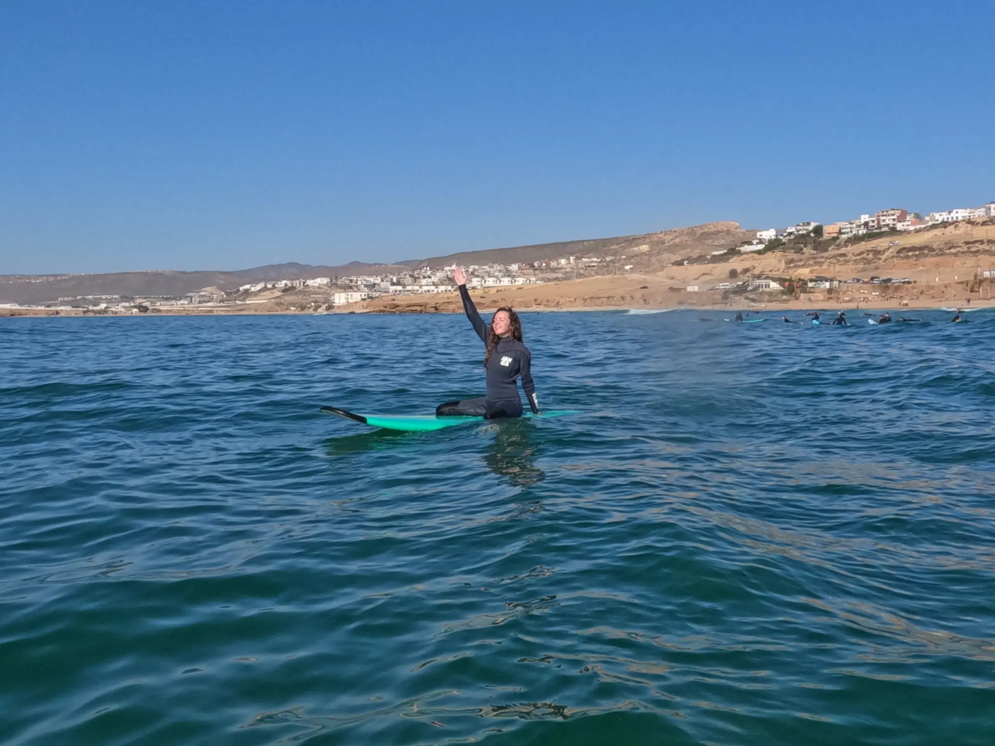 Girl sitting on her surfboard with hands up in the air, smiling in the water at Taghazout.