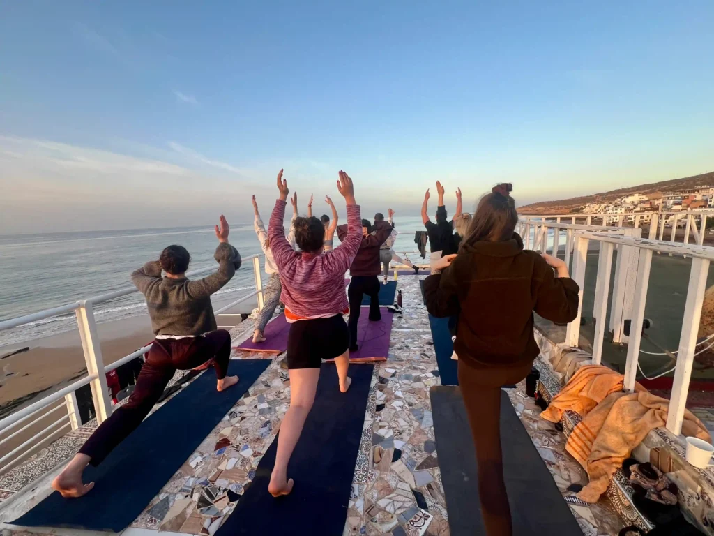 People doing yoga on a rooftop terrace in Taghazout, Morocco, with the ocean in the background at sunrise.