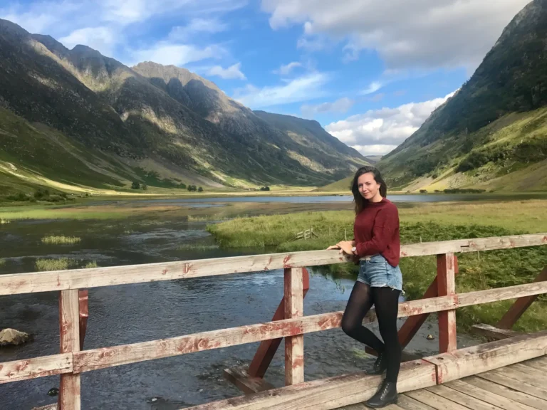 Girl standing on a bridge in Glencoe with river and mountains in the background.